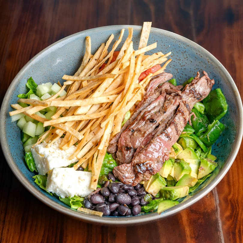 Mexican salad with grilled steak, tortilla strips, black beans, avocado, and cheese in a blue bowl.