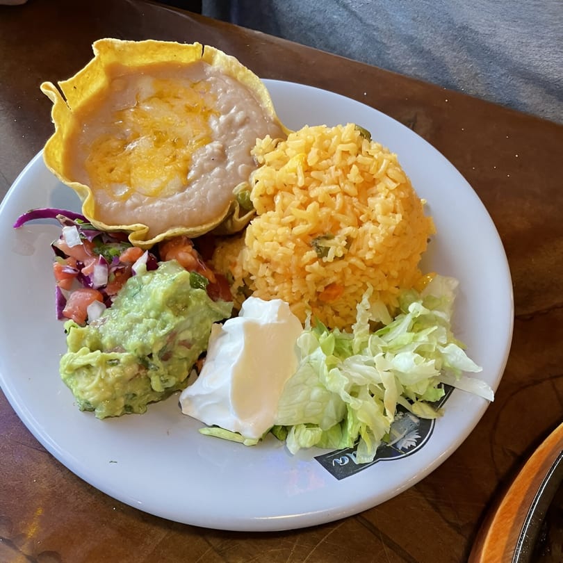 Mexican meal with refried beans in a tortilla bowl, yellow rice, guacamole, pico de gallo, sour cream, and lettuce.