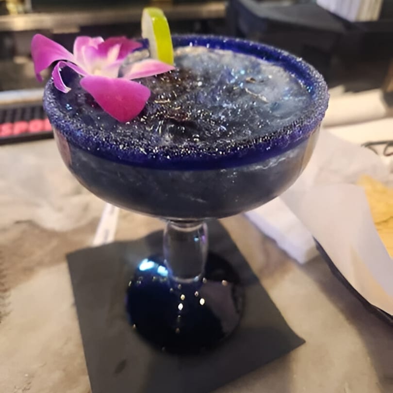 A vibrant blue margarita in a salt-rimmed glass, garnished with a lime wedge and purple orchid.