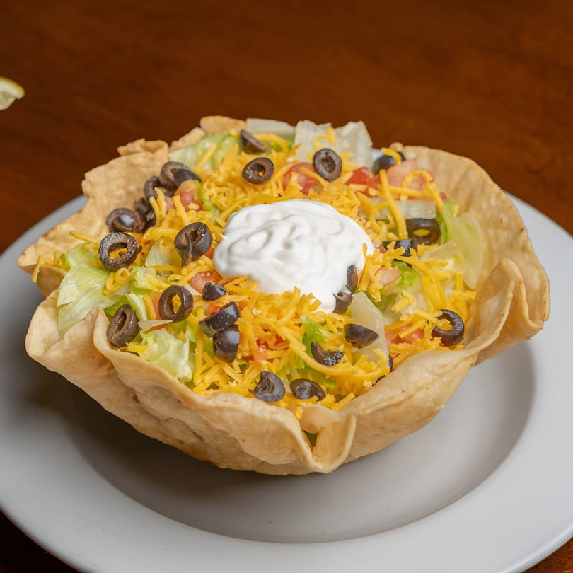A taco salad in a crispy tortilla bowl, topped with lettuce, cheese, olives, and sour cream.