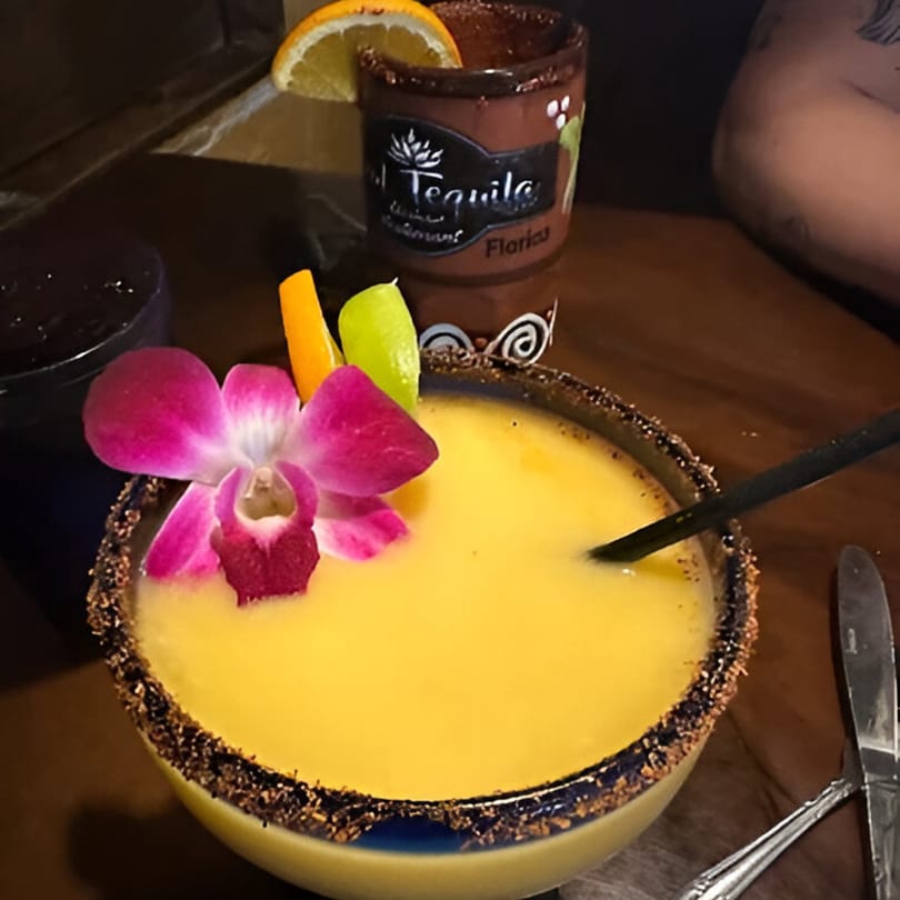 A vibrant yellow mango margarita with a pink orchid, orange slice, and lime wedge.