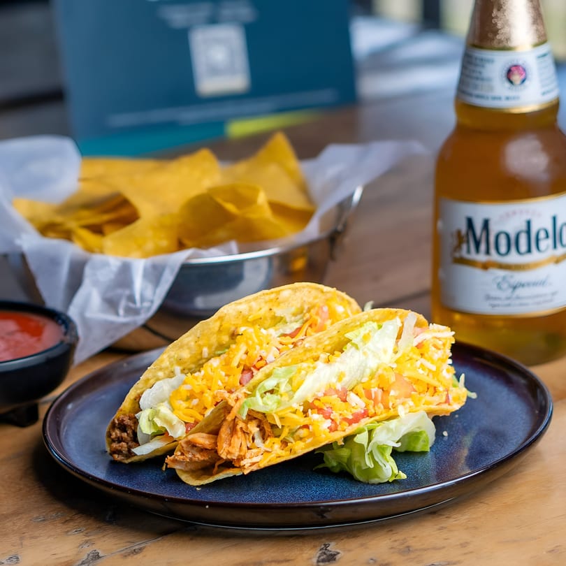 Two hard-shell tacos with meat, cheese, lettuce, and tomatoes, with chips and beer.